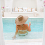 woman relaxing in resort in Europe poolside wearing a hat and holding a drink.